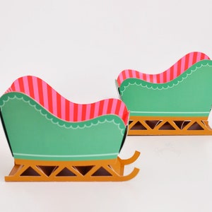 May include: Two miniature green and gold sleighs with pink and red striped seats. The sleighs are made of paper and are perfect for a Christmas craft project.