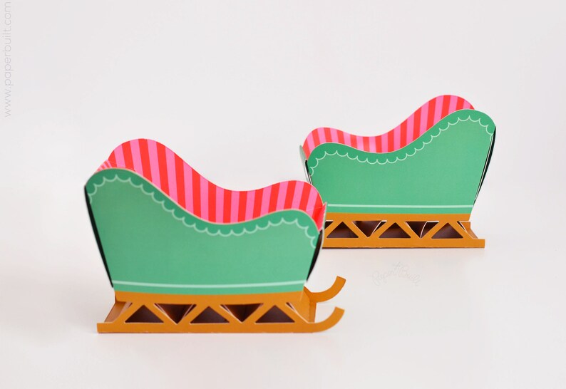 May include: Two miniature green and gold sleighs with pink and red striped seats. The sleighs are made of paper and are perfect for a Christmas craft project.