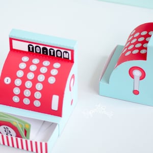 May include: A red and blue toy cash register with a drawer that opens to reveal play money. The cash register has a display that reads "TO:TOM" and a number pad with red buttons.