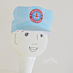 May include: A blue and white striped paper hat with a red and white circle that says "Chaseville Railroad" on it. The hat is on top of a white cone with a drawn face.