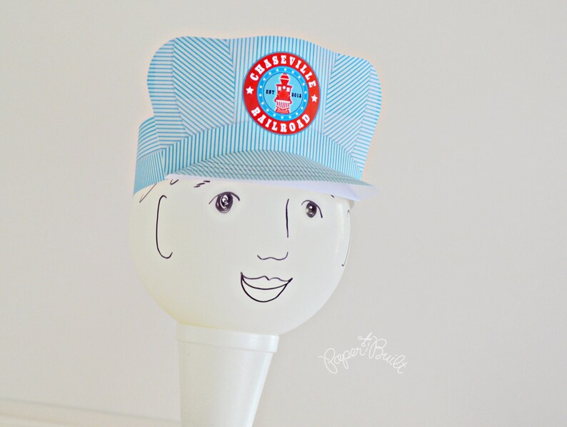 May include: A blue and white striped paper hat with a red and white circle that says "Chaseville Railroad" on it. The hat is on top of a white cone with a drawn face.