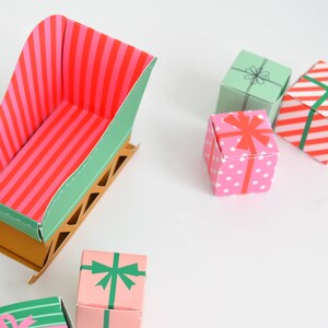 May include: A green and brown paper sleigh with a pink and red striped interior. There are five small gift boxes with various patterns, including polka dots, stripes, and a green bow.