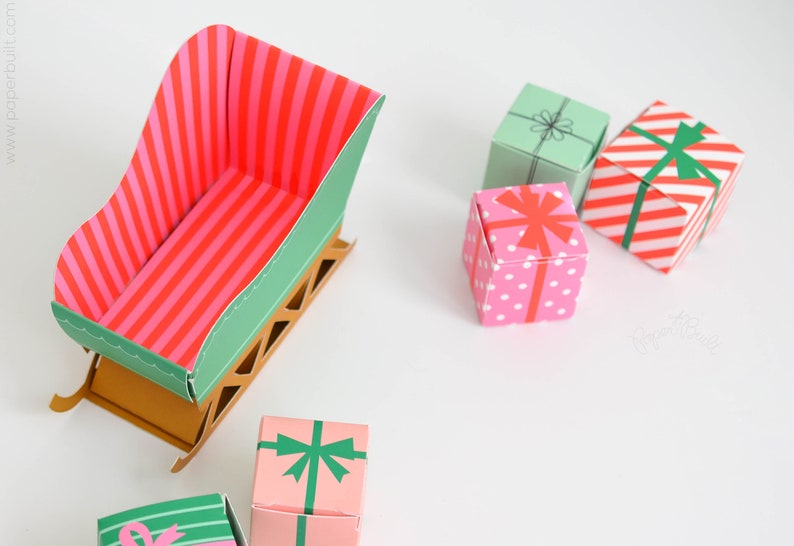 May include: A green and brown paper sleigh with a pink and red striped interior. There are five small gift boxes with various patterns, including polka dots, stripes, and a green bow.
