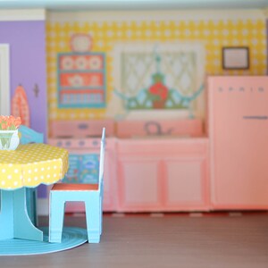 May include: A dollhouse kitchen set with a blue and yellow table and chairs, a pink refrigerator, and a yellow and white patterned wall.