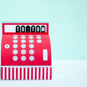 May include: Red and white papercraft cash register with the text "TO:TOM" on the top. The cash register has a number pad with numbers 1-10, 20, 30, 40, 50, 60, 70, and 80.