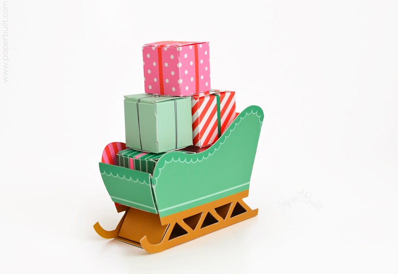 May include: A green paper sleigh with a gold base, carrying four wrapped gifts. The gifts are decorated with red and white stripes, pink polka dots, and green stripes.