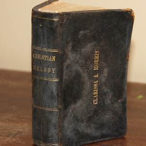 Op de afbeelding: Een zwart, versleten boek getiteld "Christian Melody" door Clarissa A. Morris.