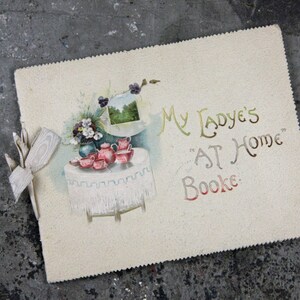 May include: A vintage book cover with a decorative illustration of a table setting with teacups and flowers. The text on the cover reads "My Ladye's "AT Home" Booke."