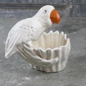 Puede incluir: Una maceta de cerámica blanca con forma de loro y pico naranja. El ave está posada sobre un cuenco con borde festoneado, con detalles verdes en las alas. Esta pieza decorativa es adecuada para plantas pequeñas o como adorno único.