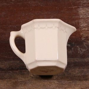 Puede incluir: Una jarra de crema de cerámica blanca con un borde festoneado y un asa. La jarra es octogonal y tiene un acabado liso y brillante.