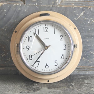 May include: A round, cream-colored Citizen clock with a white face and black numerals. The clock has a silver-colored bezel and black hands. The clock is mounted on a wall and has a black knob at the top.
