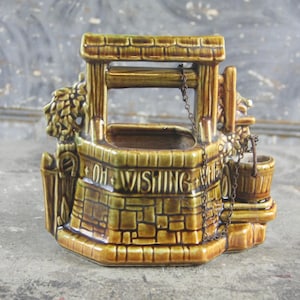 Può includere: Una fioriera a forma di pozzo dei desideri in ceramica, marrone e verde. Il pozzo presenta un design a forma di mattone, un piccolo secchio e la scritta "OH WISHING WELL". Ha un tetto e una catena.