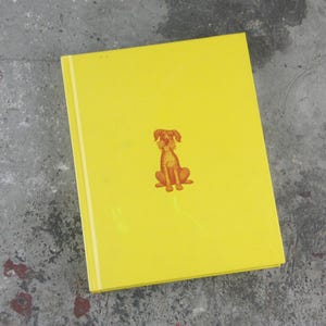 May include: A yellow book featuring a small illustration of a brown dog. The dog is depicted in a seated position. The book is closed, with a white spine visible.