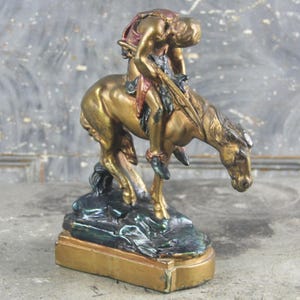 May include: A gold-toned sculpture of a person riding a horse, with the figure leaning forward over the horse's neck. The horse is positioned on a dark, textured base, and the entire piece features metallic gold highlights with dark accents.
