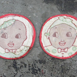 May include: Two round, red-trimmed fabric coasters with embroidered images of babies. The babies have brown skin, green eyes, and are wearing white bonnets with green bows.