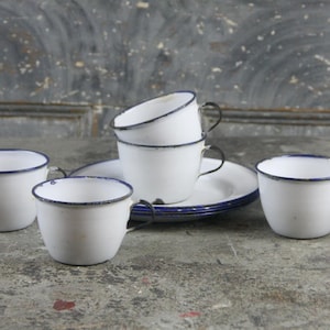 May include: Set of five white enamel mugs with blue trim and black handles. The mugs are stacked on top of each other and are sitting on a white enamel saucer.