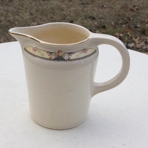 May include: A white ceramic jug with a gold rim and floral design. The jug has a handle and is decorated with pink and blue flowers.
