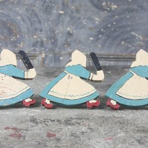 May include: Three vintage wooden figures of women in blue dresses, white aprons, and red shoes. Each figure holds a black object. The figures are painted in a folk art style and are set against a gray background.