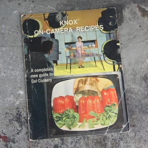 以下が含まれることがあります： レトロなデザインの「KNOX ON-CAMERA RECIPES」というタイトルのヴィンテージ料理本。表紙には、キッチンにいる女性と、ゼラチン料理を表示するテレビ画面が描かれています。「A completely new guide to Gel-Cookery」というテキストも表示されています。