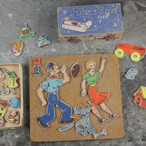 May include: A vintage wooden puzzle featuring a cartoon couple, a dog, a house, and other objects. The puzzle pieces are colorful and have a retro style. The box for the puzzle is blue and white and says "Rose Bud Sled".