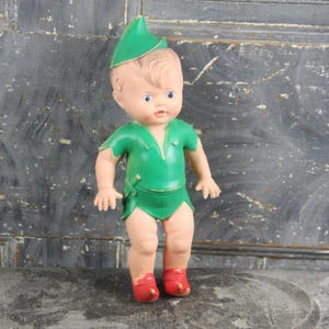 May include: Vintage doll dressed in a green tunic and hat, with red shoes. The doll has blonde hair and blue eyes. The doll's outfit is reminiscent of a storybook character. The doll is standing upright.