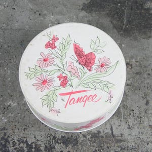 May include: A round, white vintage tin with a floral and butterfly design. Pink flowers and a pink butterfly are featured, along with green leaves. The word "Tangee" is written in pink script across the bottom.