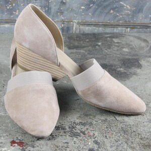 May include: A pair of beige suede wedge sandals. The open-sided shoes have a pointed toe and a low wedge heel. A beige elastic band stretches across the top of the foot. The shoes are displayed on a gray surface.