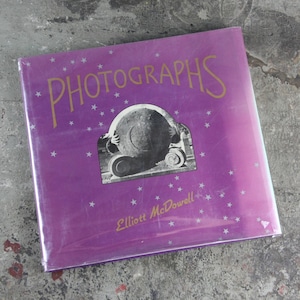 May include: A purple book with a gold title "PHOTOGRAPHS" and the author's name "Elliott McDowell" printed on the cover. The cover features a black and white photo of a large stone sphere.