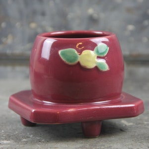 May include: A burgundy ceramic planter with a square base and three small feet. The planter has a round top and is decorated with a floral design in yellow and green. The planter is likely for small plants or succulents.