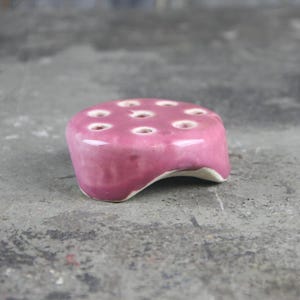 May include: A small, pink ceramic flower frog with multiple holes for holding stems. The frog has a rounded, slightly curved base.