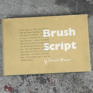 May include: A rectangular book with a tan cover featuring the white text Brush Script and the author name by Donald Stevens. The cover includes a paragraph of black text discussing the history and utility of brush script lettering in commercial art.