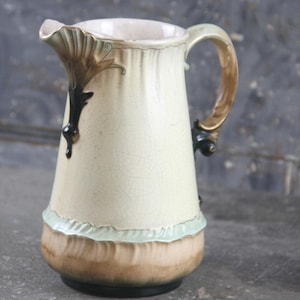 Puede incluir: Jarra de cerámica color crema con borde y asa con volantes. La jarra presenta detalles florales marrones y negros cerca del pico y el asa, con un ribete verde claro en la base. La base es marrón y el asa es dorada.