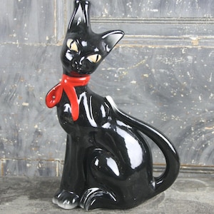 Puede incluir: Una figura de gato de cerámica negra brillante con una bufanda roja brillante atada al cuello. El gato tiene orejas puntiagudas, ojos amarillos almendrados y una cola larga y curva. La superficie de la cerámica es lisa y reflectante, destacando la postura erguida y alerta del gato y sus rasgos faciales estilizados.