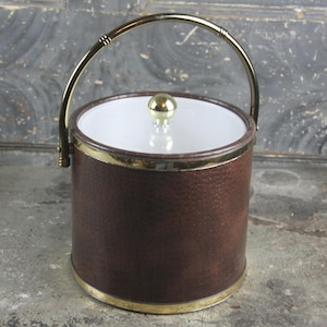 May include: A brown faux leather ice bucket with a gold-toned handle and lid. The lid has a gold-toned knob.