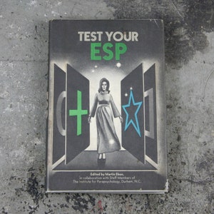 Puede incluir: Un libro titulado "Test Your ESP" con una mujer con un vestido blanco de pie frente a tres puertas. Las puertas tienen símbolos en ellas: un círculo, una cruz y una estrella.