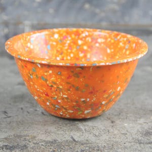 May include: A round bowl with a vibrant orange base color featuring a multi-colored confetti or terrazzo-style pattern throughout the exterior and interior surfaces. The bowl has a smooth, glossy finish and a wide, circular rim with a curved edge.