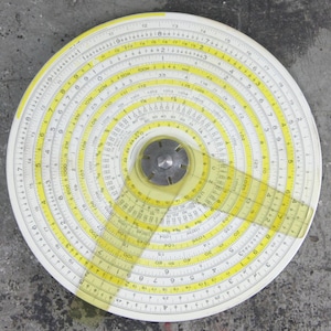 May include: A circular slide rule with white and yellow markings. The slide rule has multiple concentric rings with numbers and scales. A clear plastic arm with yellow markings pivots from the center.