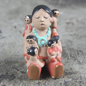 May include: A tan clay figurine depicting a central figure with closed eyes and a turquoise-colored necklace, surrounded by several smaller figures attached to the body. The figures have black hair and are wearing red clothing accents on a tan base.