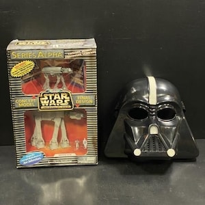 May include: A black Darth Vader mask with a white stripe and a Star Wars Series Alpha model box. The box features a model of a Star Wars vehicle and the text "CONCEPT MODEL" and "FINAL DESIGN".