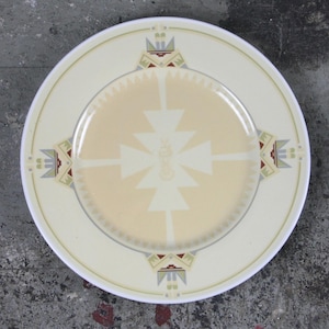 May include: Cream-colored ceramic plate with a geometric design. The plate features a central pattern of cream and beige triangles, surrounded by a border with repeating geometric shapes in beige, green, and red. The plate has a diameter of approximately 10 inches.