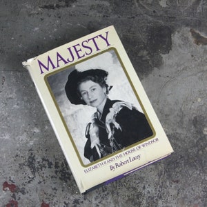 May include: A white book with the title "Majesty: Elizabeth II and the House of Windsor" by Robert Lacey. The cover features a black and white photo of Queen Elizabeth II wearing a hat and a fur collar.