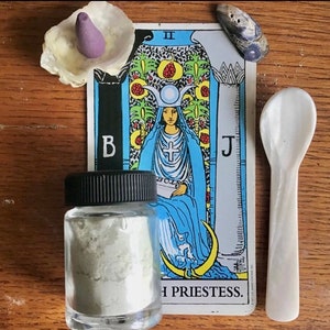 May include: A tarot card reading with the High Priestess card, a small jar of white powder, a white ceramic spoon, and a seashell with a purple incense cone.