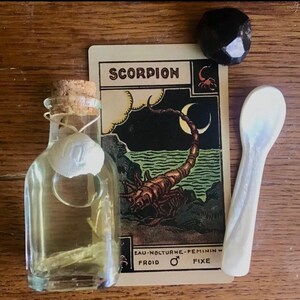 Puede incluir: Una carta de tarot con la palabra "SCORPION" impresa en ella, una pequeña botella de vidrio con un tapón de corcho, una concha de mar blanca y una cuchara de cerámica blanca. La carta de tarot tiene una ilustración de un escorpión y las palabras "EAU-NOCTURNE-FEMININ-FROID-FIXE".