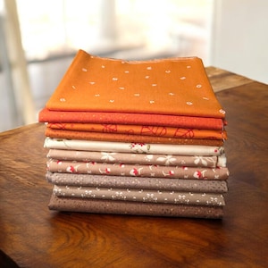 Puede incluir: Una pila de cuadrados de tela doblados en varios colores y patrones. La capa superior es naranja con estampados de letras y números blancos. Otras telas incluyen diseños florales y de lunares en tonos de marrón, beige y rojo. Las telas están cuidadosamente apiladas sobre una mesa de madera.