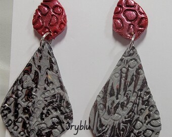 May include: A pair of dangle earrings with a red and silver design. The top of the earrings are a small red teardrop shape with a textured surface. The bottom of the earrings are a larger silver teardrop shape with a textured surface. The earrings are handmade with love.