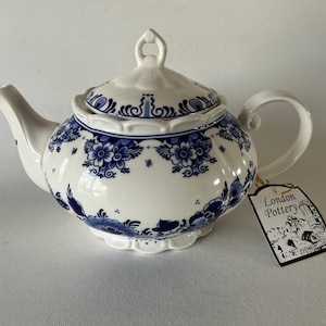 May include: White ceramic teapot with blue floral patterns and a matching lid. The teapot has a curved handle and spout. A tag that says "London Pottery" is attached.