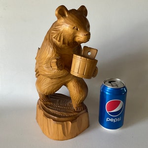 May include: A wooden carving of a bear holding a bucket. The bear is standing on a wooden base. The carving is a light brown color.