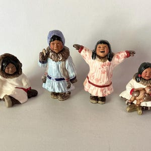 May include: Four ceramic figurines depicting children in winter clothing. One sits in a white parka, one stands in a blue striped parka with a hood, one stands with arms outstretched in a pink floral parka, and one sits holding a small brown teddy bear.