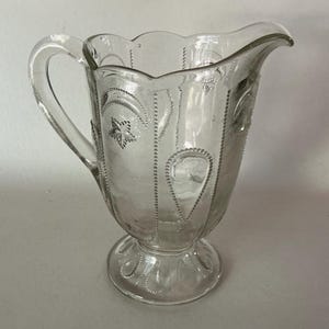 May include: A clear glass pitcher featuring a scalloped rim, a curved handle, and a pedestal base. The exterior surface is decorated with embossed patterns including stars, crescent moons, and teardrop shapes outlined with beaded detailing.