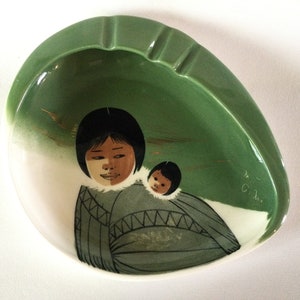 May include: Green and white ceramic ashtray with a painted image of a woman and child in traditional clothing. The ashtray is oval shaped with a raised rim.